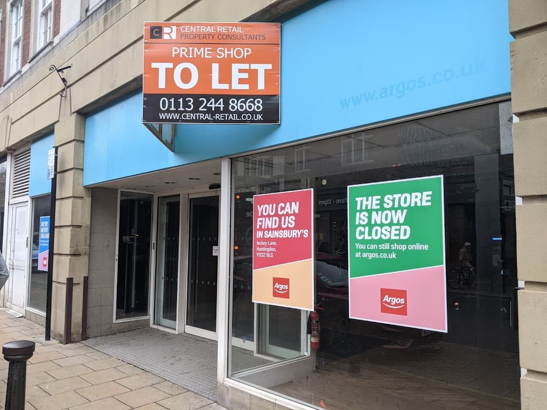 Closed storefront with "TO LET" sign and notices for Sainsbury's and Argos, featuring large glass windows.