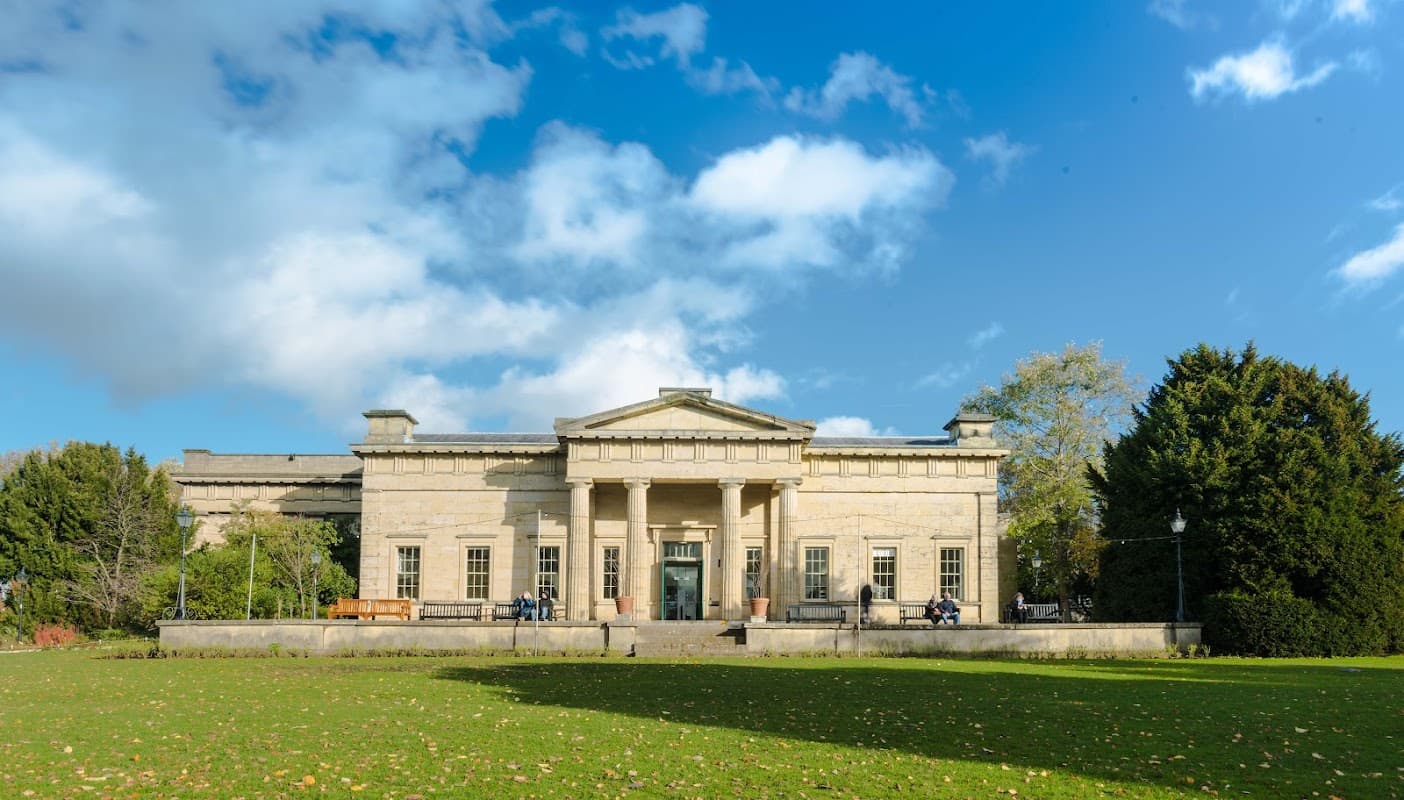 Neoclassical building with columns, surrounded by green grass and trees under a blue sky with scattered clouds.