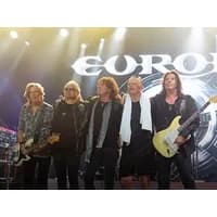 Band Europe on stage, five members with instruments, bright stage lights, large logo backdrop.