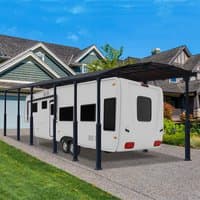 Grey metal carport with arched roof, supporting poles, and open sides, covering a large vehicle in a driveway.