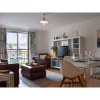 Modern living room with brown leather sofa, white dining table, TV on white shelf, and nautical decor by large window.