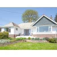 White and blue single-story cottage with large windows, sloped roof, and a manicured lawn under a clear sky.