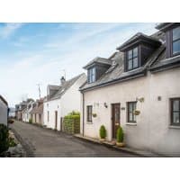 White seaside cottage with dormer windows, wooden door, and potted plants, part of "By the Sea" by cottages.com.