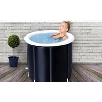 Black portable hot tub with white rim, filled with water, person relaxing inside, placed on wooden deck near potted plant.