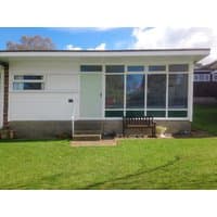 White holiday cottage with large front windows, green door, and wooden bench on a grassy lawn.