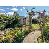 Cottage with blue exterior, surrounded by lush garden, stone path, and wooden archway under a clear blue sky.