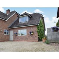 Red brick cottage with pitched roof, large front window, hanging flower baskets, and gravel driveway.
