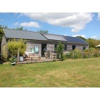 "The Cowshed" cottage with wooden exterior, solar panels, and lush garden under a blue sky.