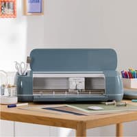 Cricut Maker 4 Electronic cutting machine, gray with open lid, on wooden table with crafting supplies nearby.