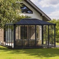 Black-framed enclosed gazebo with clear glass panels and a sloped roof, set in a grassy yard near a brick house.