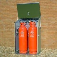 Green metal gas storage unit with open lid, housing two large red gas cylinders, against a brick wall.