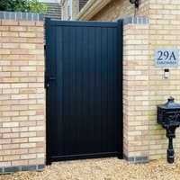 Black Devon Premium Metal Side Gate with vertical panels, sleek design, and sturdy frame, set between brick pillars.
