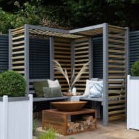 Corner garden arbour seat with grey frame, slatted roof and sides, wooden bench, and decorative cushions.