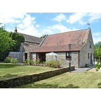 Stone cottage with red-tiled roof, chimney, and garden. Surrounded by a low stone wall and greenery under a blue sky.
