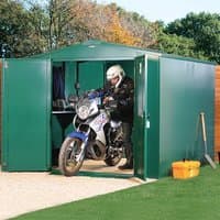 Green metal Asgard motorbike garage with wide double doors, housing a motorcycle, set on a gravel base.