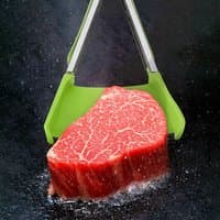 Green and silver Avilo 2 in 1 Spatula & Tongs holding a raw steak, featuring a wide silicone grip and stainless steel arms.