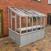 Green-painted wooden lean-to greenhouse with clear panels, sloped roof, and single door, set against a brick wall.