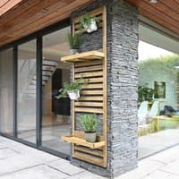 Tall wooden wall planter with slatted design, two shelves, and potted plants, mounted on a stone exterior wall.