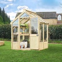 Wooden greenhouse with clear panels, pitched roof, and door, set in a grassy garden with trees and a house nearby.