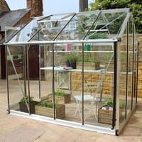 Green metal-framed greenhouse with toughened glass panels, pitched roof, and sliding door, housing plants and a chair.