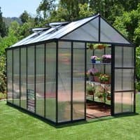 Black-framed 8x12 Palram Canopia Glory Greenhouse with clear panels and roof vents, set on grass with plants inside.