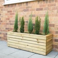 Wooden garden planter, 4'x1', with horizontal slats, filled with tall green plants against a brick wall.