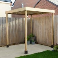 Wooden garden pergola with square frame, slatted roof, and four sturdy posts on a patio near a wooden fence.