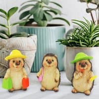 Set of 3 hedgehog figurines with hats and bags, each in a unique pose, surrounded by potted plants.